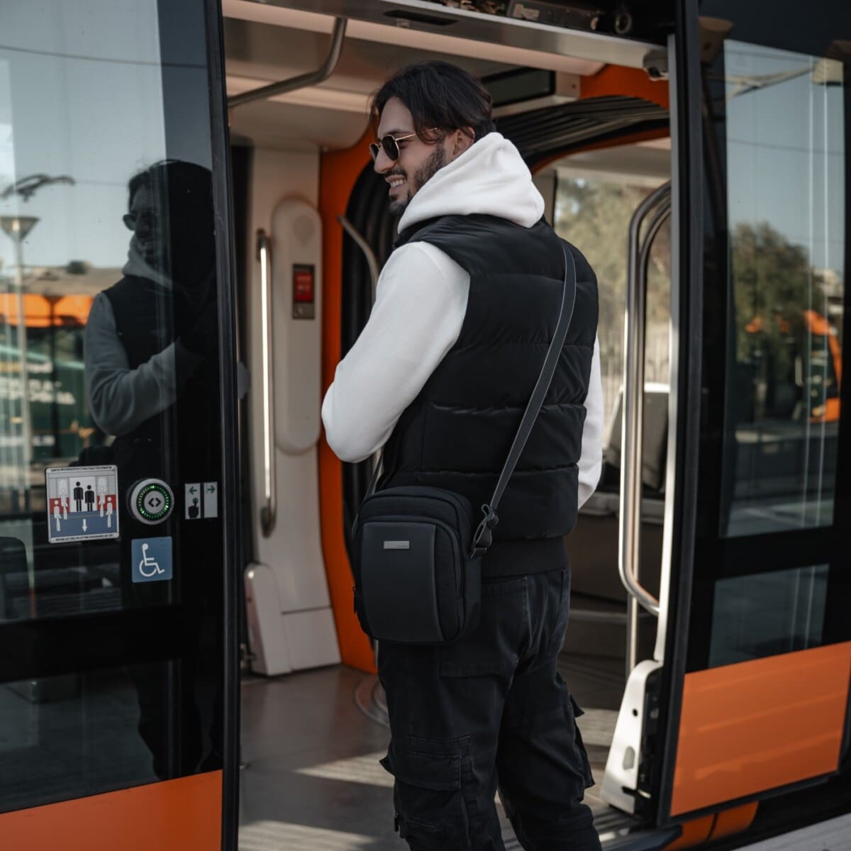 Φωτογραφία ενός άνδρα με casual ντύσιμο να βγαίνει από ένα τραμ. Φοράει λευκό φούτερ με κουκούλα, μαύρο γιλέκο, σκούρο παντελόνι και γυαλιά ηλίου. Κρατάει μια μαύρη τσάντα χιαστί cardinal . Το τραμ είναι πορτοκαλί και λευκό, και στο βάθος φαίνονται άλλοι άνθρωποι και ένας στύλος φωτισμού. Ο άνδρας χαμογελάει.