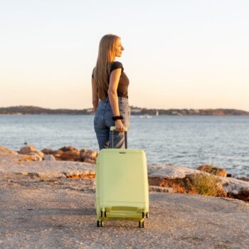 Γυναίκα με μαύρο μπλουζάκι και τζιν προχωράει με κίτρινη μεσαία βαλίτσα σε παραλία, με τη θάλασσα και τον ορίζοντα στο φόντο.